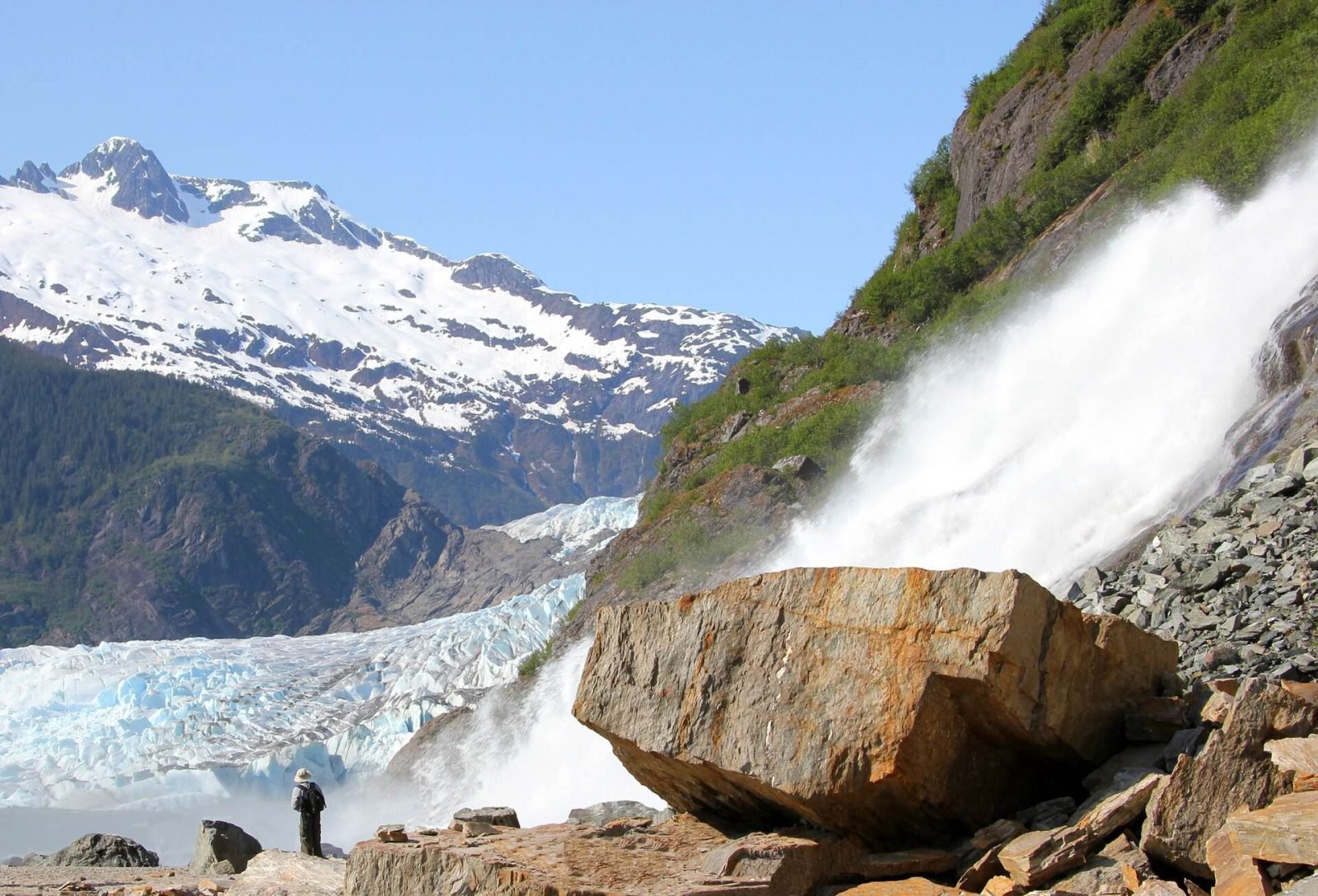 Mendenhall Glacier beside Nugget Falls under a clear blue sky, with a visitor standing on the rocks