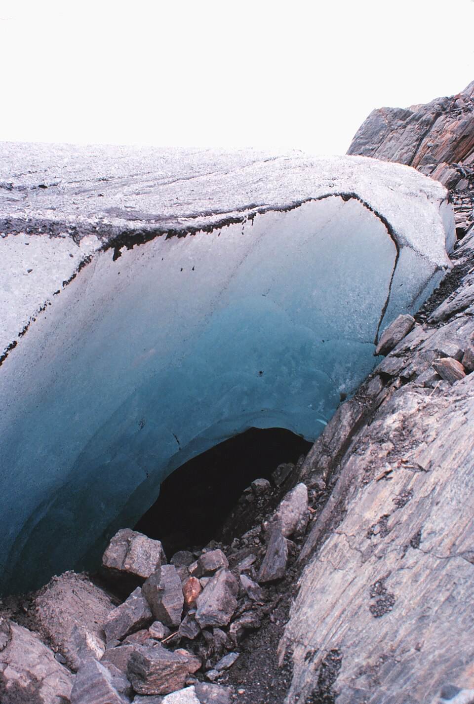 The dark entrance to an ice cave beneath a curved wall of blue glacier ice