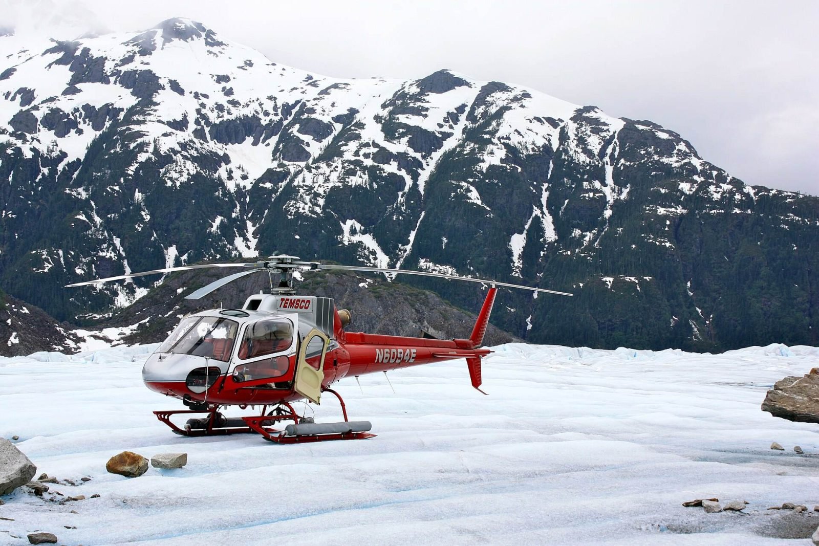 A red helicopter parked on the snow of Mendenhall Glacier below jagged peaks