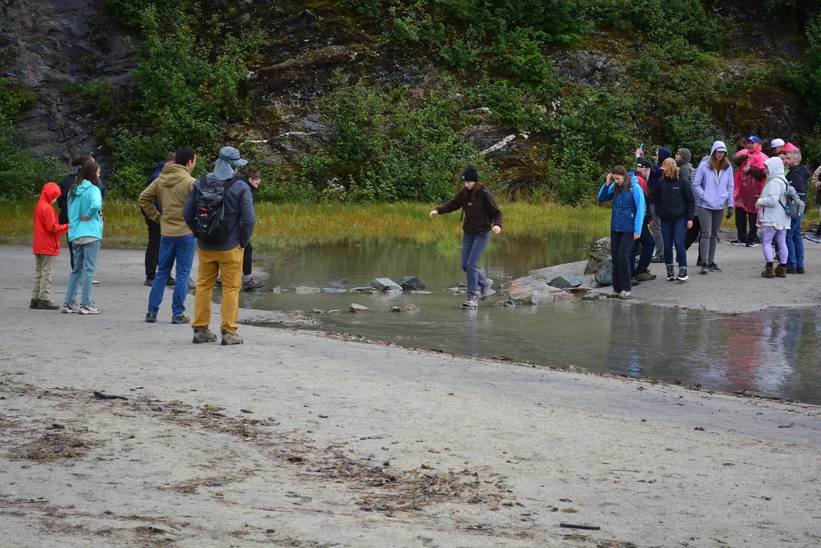 Visitors in rain jackets and hiking shoes crossing stepping stones on the gravel beach near Nugget Falls